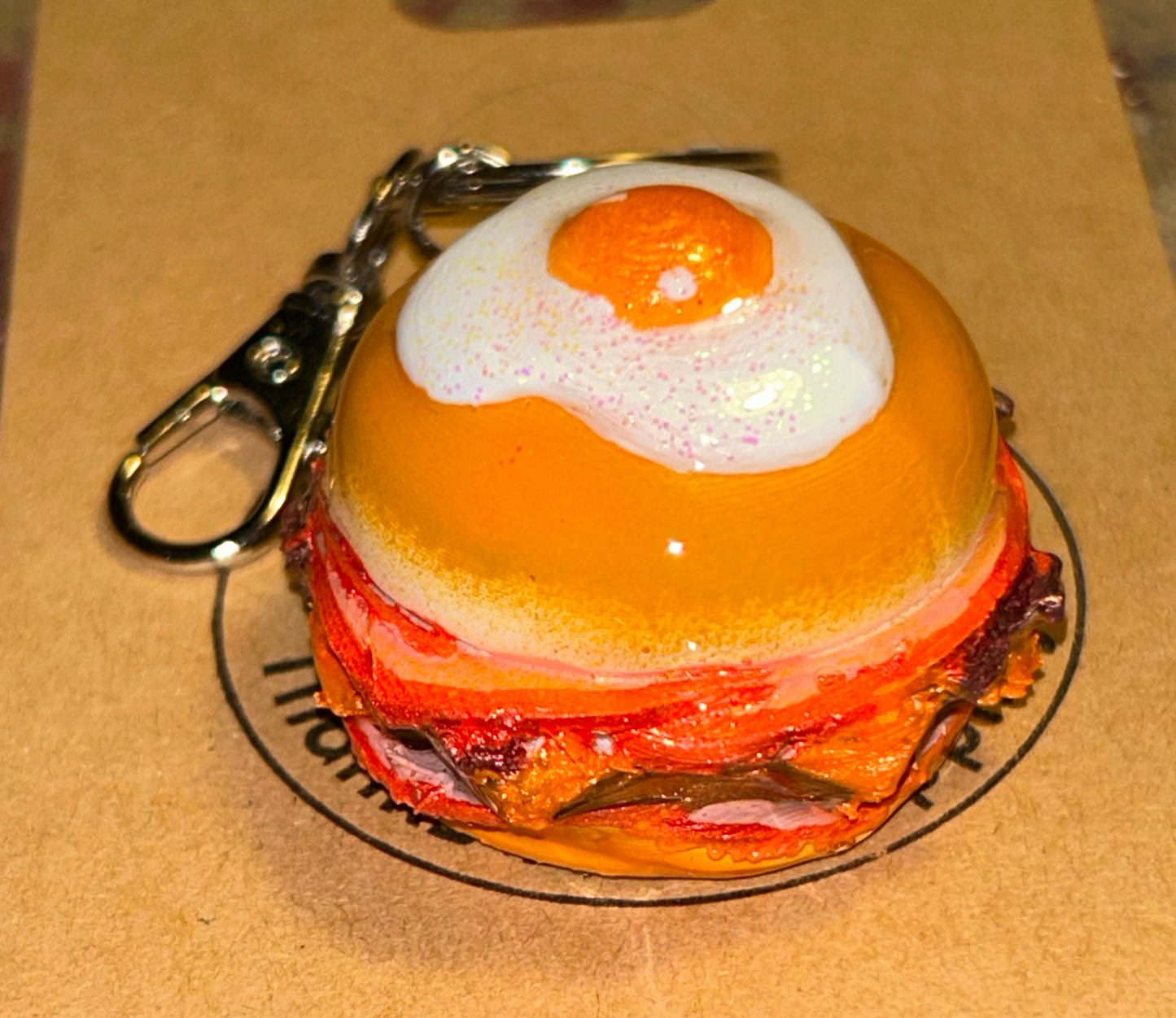 Keychain shaped like a breakfast burger with a bun, meat, and egg on a cardboard background
