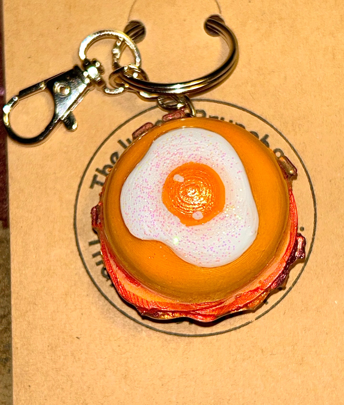 Keychain shaped like a breakfast burger with a bun, meat, and egg on a cardboard background