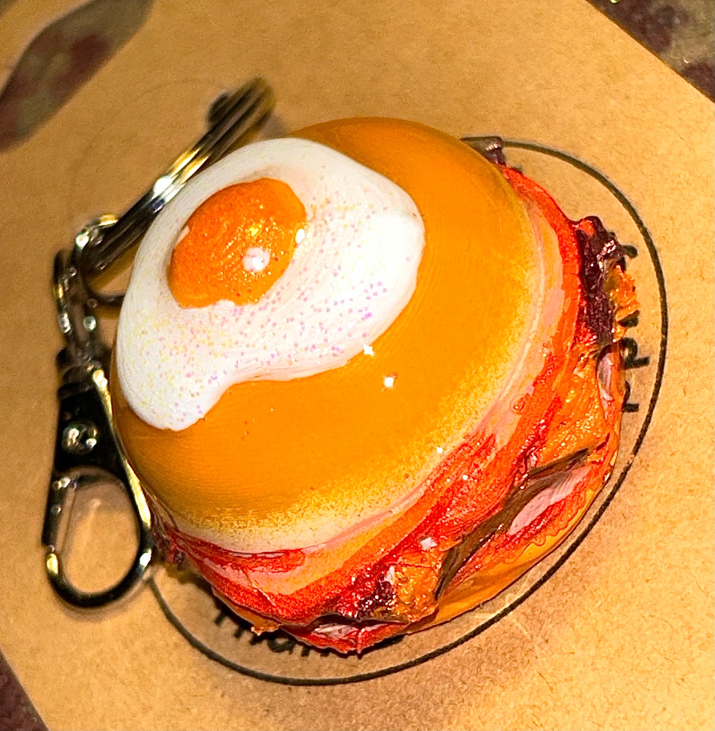 Keychain shaped like a breakfast burger with a bun, meat, and egg on a cardboard background